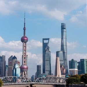 Lo skyline moderno di Shanghai, con la Torre della Perla Orientale, si erge sopra un fiume e un ponte d'acciaio sotto un cielo blu con nuvole bianche.