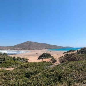 Una vista panoramica di un'ampia spiaggia sabbiosa con acqua turchese e onde, vista da una collina coperta da arbusti verdi sotto un cielo azzurro chiaro.