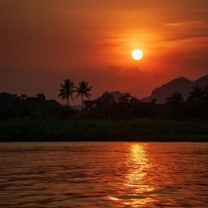 Il sole tramonta in un cielo arancione intenso, la sua luce si riflette su un fiume con sagome di palme e colline sulla riva opposta.