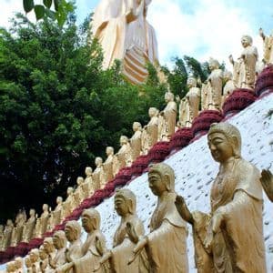 Eine riesige goldene Buddha-Statue thront über vielen kleineren goldenen Statuen, die in Reihen an einem Berghang unter einem teilweise bewölkten Himmel angeordnet sind.
