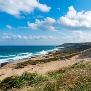 Eine sandige Küstenlinie schlängelt sich entlang eines blauen Ozeans mit sanften Wellen, von einem grasbewachsenen Hügel unter einem hellen, teils bewölkten Himmel aus gesehen.