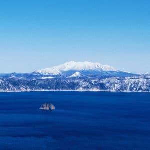 Un'ampia vista di un lago blu profondo con un piccolo isolotto roccioso, sullo sfondo montagne innevate sotto un cielo limpido.