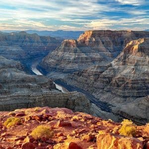 Un vasto canyon stratificato con un fiume sottostante, ammirato da un promontorio roccioso rosso all'alba, sotto un cielo parzialmente nuvoloso.