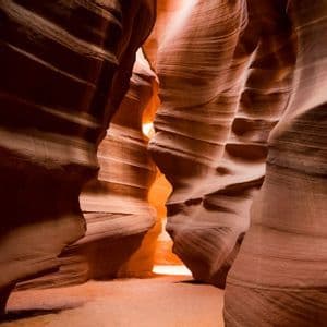 La luce del sole illumina lo stretto passaggio di un canyon a fessura, mettendo in risalto le pareti fluide di arenaria arancione e rossa.