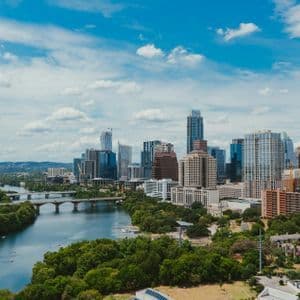 Uno skyline urbano con grattacieli si staglia dietro un ampio fiume, circondato da una vegetazione lussureggiante, sotto un cielo parzialmente nuvoloso.