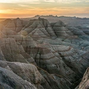Un vasto paesaggio di formazioni rocciose stratificate (calanchi) illuminato dalla calda luce di un tramonto sotto un cielo parzialmente nuvoloso.