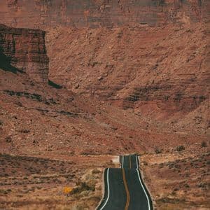 Una strada asfaltata vuota con linee gialle si snoda su e giù attraverso un paesaggio arido verso un'imponente parete di canyon roccioso rosso.
