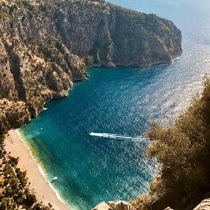 Vista dall'alto di un motoscafo che solca l'acqua turchese di una cala, con una spiaggia sabbiosa adagiata ai piedi di una grande scogliera.