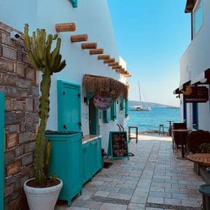 Uno stretto vicolo in pietra tra edifici imbiancati con dettagli turchesi conduce a un mare blu con barche all'orizzonte.