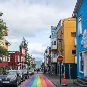 Una strada dipinta con un arcobaleno conduce verso il mare, fiancheggiata da edifici colorati e auto parcheggiate sotto un cielo nuvoloso.