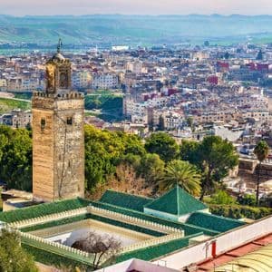 Un minarete de piedra y una mezquita con tejado de tejas verdes se alzan sobre un denso paisaje urbano, con colinas verdes al fondo.