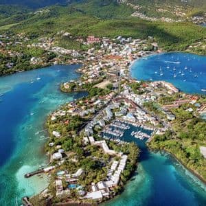 Vue aérienne d'une ville côtière sur une presqu'île, dotée d'un port de plaisance rempli de bateaux, entourée d'eaux turquoise et de collines verdoyantes.