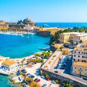 Vista aerea di una storica città costiera con una fortezza su una collina, un porto pieno di barche a vela e acque blu intenso sotto un cielo sereno.