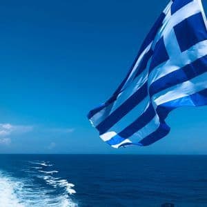 La bandiera greca sventola al vento dalla poppa di una barca, con il mare blu profondo e la scia dell'imbarcazione.