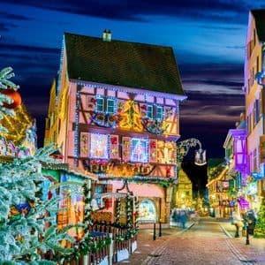 A cobblestone street at dusk lined with buildings and a Christmas tree, all illuminated with festive lights and decorations.