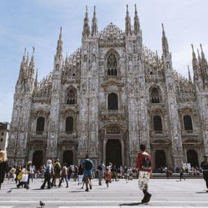 La ornamentada fachada de mármol blanco de una gran catedral gótica vista desde una animada plaza pública llena de gente en un día soleado.