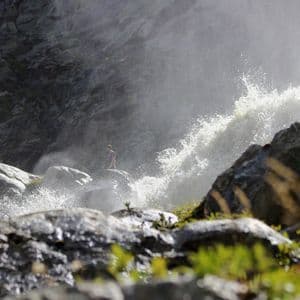 Eine Person steht auf einem großen Felsen neben dem gewaltigen Sprühnebel und der Gischt eines rauschenden Wasserfalls.