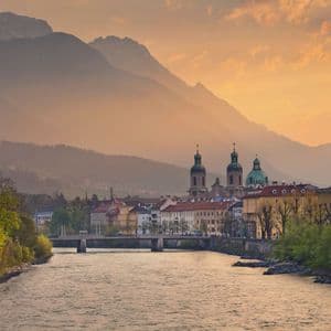 Eine historische Stadt mit markanten Kirchtürmen am Flussufer, vor der Kulisse hoch aufragender, diesiger Berge bei Sonnenuntergang.