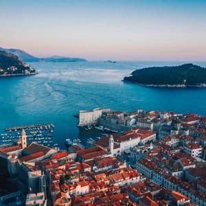 Una vista aerea di una storica città costiera con tetti rossi, che si affaccia su un mare blu profondo e un'isola in lontananza al tramonto.