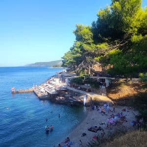Una vista dall'alto di persone che nuotano e prendono il sole su una spiaggia di ciottoli, accanto a un caffè su una scogliera alberata.