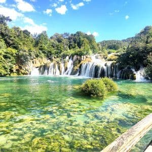 Una cascata a più livelli si riversa in un limpido fiume turchese, circondata da una rigogliosa vegetazione sotto un cielo azzurro e soleggiato.