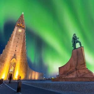 Eine moderne Kirche und eine Statue stehen nachts unter dem leuchtenden grünen Nordlicht beleuchtet.