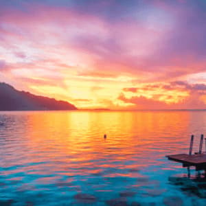 Un bungalow sull'acqua con tetto di paglia si affaccia su un mare calmo sotto un tramonto rosa e arancione vibrante, con montagne in lontananza.