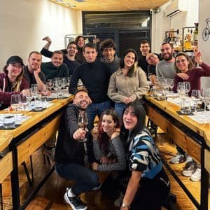 Un gruppo WeRoad posa per una foto intorno a lunghi tavoli di legno apparecchiati con bicchieri da vino durante un evento di degustazione al chiuso.