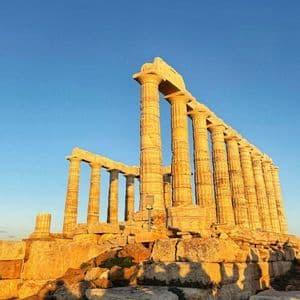 Golden sunlight illuminates the stone columns of an ancient temple ruin against a clear blue sky.