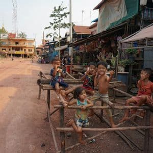 Un gruppo di bambini sorride e posa per la macchina fotografica, seduti su una struttura di legno lungo una strada sterrata in un villaggio.