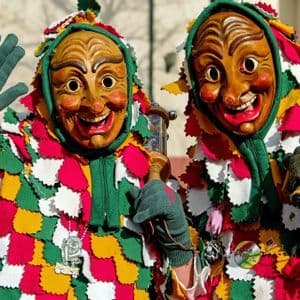 Due persone in costumi variopinti a patchwork e grandi maschere di legno sorridenti, con una che saluta.
