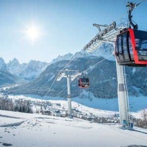 Teleféricos rojos y negros viajan sobre una pendiente de montaña nevada en un día soleado, con una vasta cordillera al fondo.