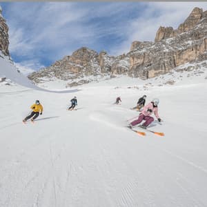 Un viaje en grupo de cinco personas de WeRoad esquiando por una amplia pista nevada rodeada de imponentes montañas rocosas bajo un cielo azul.