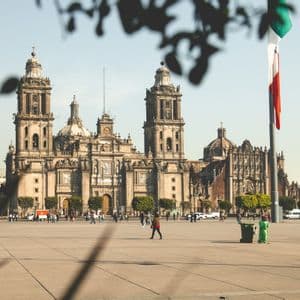 Una grande e sontuosa cattedrale con due campanili si affaccia su un'ampia piazza pubblica soleggiata, con gente che passeggia e una bandiera nazionale che sventola.