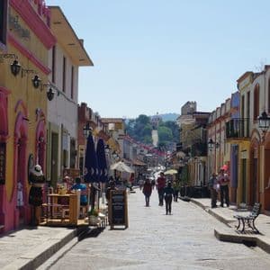 Persone passeggiano lungo una strada acciottolata e assolata, fiancheggiata da colorati edifici coloniali, con vista su una struttura su una collina lontana.