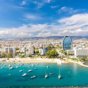 Vue aérienne d'une ville côtière moderne, avec de nombreux bateaux dans une baie turquoise et un bâtiment en verre ovale distinctif sur le littoral.