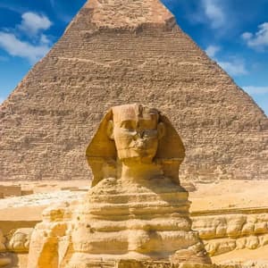 Die Große Sphinx von Gizeh steht an einem sonnigen Tag mit blauem Himmel und weißen Wolken vor einer der großen Pyramiden.