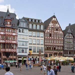 Una plaza histórica europea llena de gente, bordeada de edificios tradicionales de entramado de madera y una torre de catedral gótica al fondo.