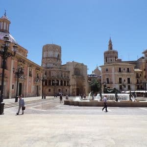 Una piazza cittadina europea illuminata dal sole, con una grande fontana, edifici storici e persone che passeggiano in una giornata serena.