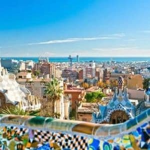 Un paesaggio urbano con edifici unici e bizzarri e una guglia blu e bianca, visto oltre un muro a mosaico colorato.