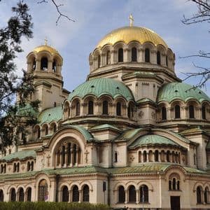 Una grande cattedrale con cupole dorate e verdi si erge sotto un cielo parzialmente nuvoloso, incorniciata da rami d'albero.