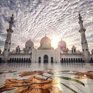 Une mosquée blanche aux dômes et minarets se reflétant sur une cour polie, ornée d'une grande mosaïque florale, sous un ciel texturé et nuageux.