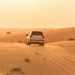 Un SUV argentato percorre vaste dune di sabbia in un paesaggio desertico durante un tramonto dorato, sollevando sabbia.
