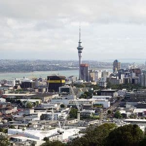 Veduta grandangolare dall'alto di uno skyline moderno della città, con un'alta torre prominente, numerosi edifici e uno specchio d'acqua in lontananza.
