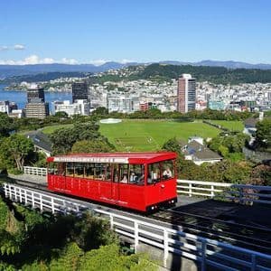 Una funivia rossa viaggia lungo un binario con una vista panoramica su una città costiera, un grande parco e montagne sullo sfondo.