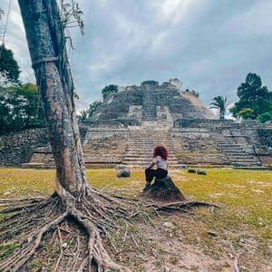 Una persona dai capelli rossi siede su una roccia ammirando un'antica piramide di pietra, con le radici di un grande albero in primo piano.