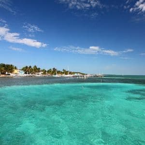 Una vista sull'acqua limpida e turchese che mostra una costa tropicale con palme, edifici colorati e una barca a vela bianca ancorata.
