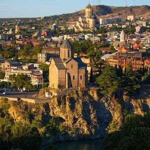 Una storica chiesa in pietra si erge su una scogliera rocciosa che domina una vasta città, con una cattedrale più grande visibile su una collina sullo sfondo.