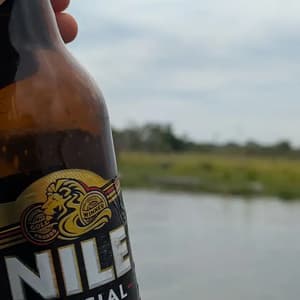 Primer plano de una mano sosteniendo una botella de cerveza Nile Special con un cuerpo de agua y un paisaje verde de fondo.