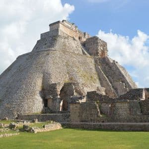 Una grande antica piramide di pietra con angoli arrotondati si erge su un campo erboso con altre rovine sotto un cielo parzialmente nuvoloso.